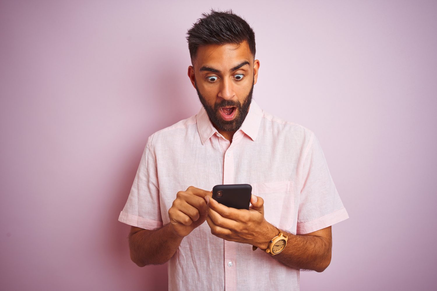 Young indian man using smartphone standing over isolated pink background scared in shock with a surprise face, afraid and excited with fear expression