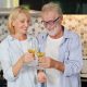 Couple senior caucasian husband and wife in casual dress standing, raising drink for cheers celebration in kitchen at house. Happy elderly man and woman holding glass of wine or cocktails with smiling