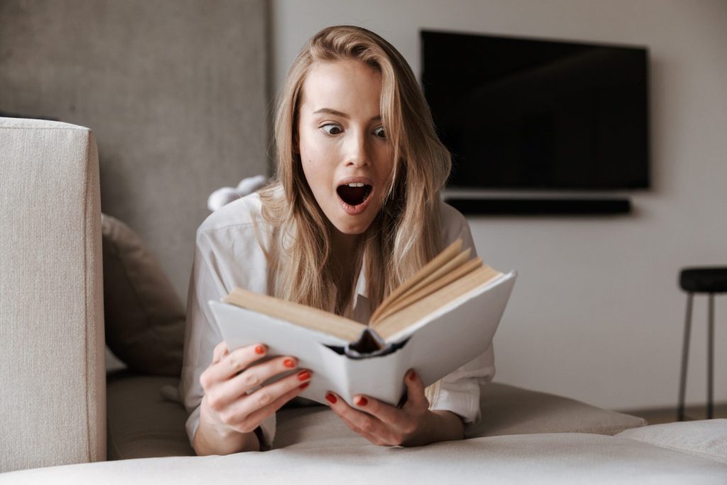 Shocked young woman holding blank cover open book while relaxing on a couch at home