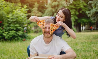 Young couple eating pizza in the park. They treat each other and laugh. Green background