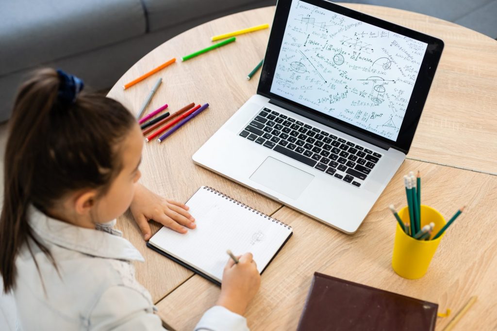 Smart little Caucasian girl child sit at table study at laptop make notes write in notebook, small schoolgirl handwrite prepare homework assignment at home, distant education concept