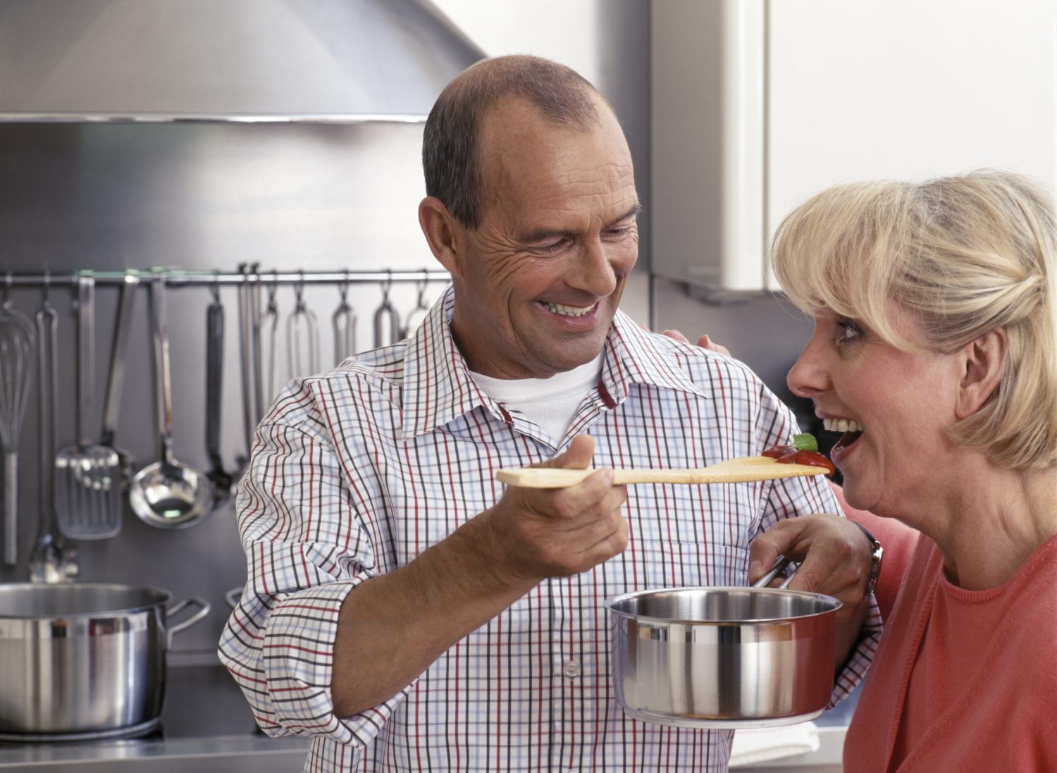 Middle-aged man feeding woman from wooden spoon in kitchen