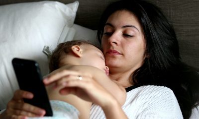 Mother with sleeping baby and checking smartphone in bed.