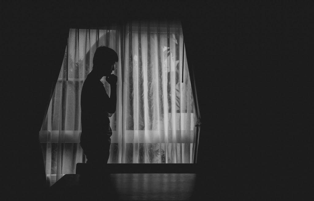 Black and white photo alone man silhouette standing with hand on chin at the window closed with curtains in darkroom. Man stands at window alone, Depression and anxiety disorder concept.