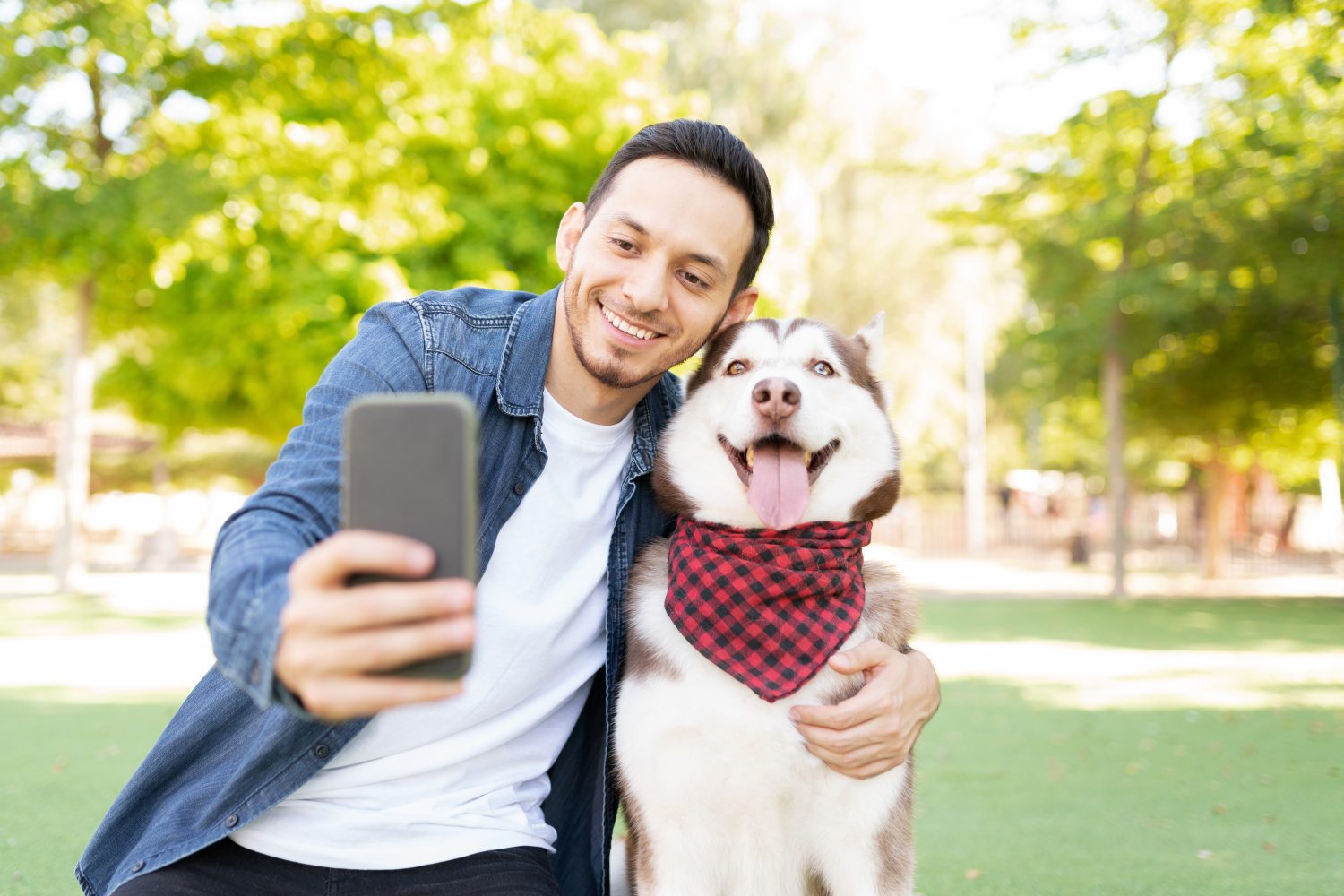 Front view of a good looking man in his 30s taking a photo with his beautiful dog wearing a bandana in the grass at the park
