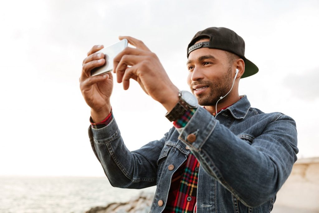 Photo of happy african man wearing cap walking on the beach while listening music with earphones. Take a photo of sea by phone.