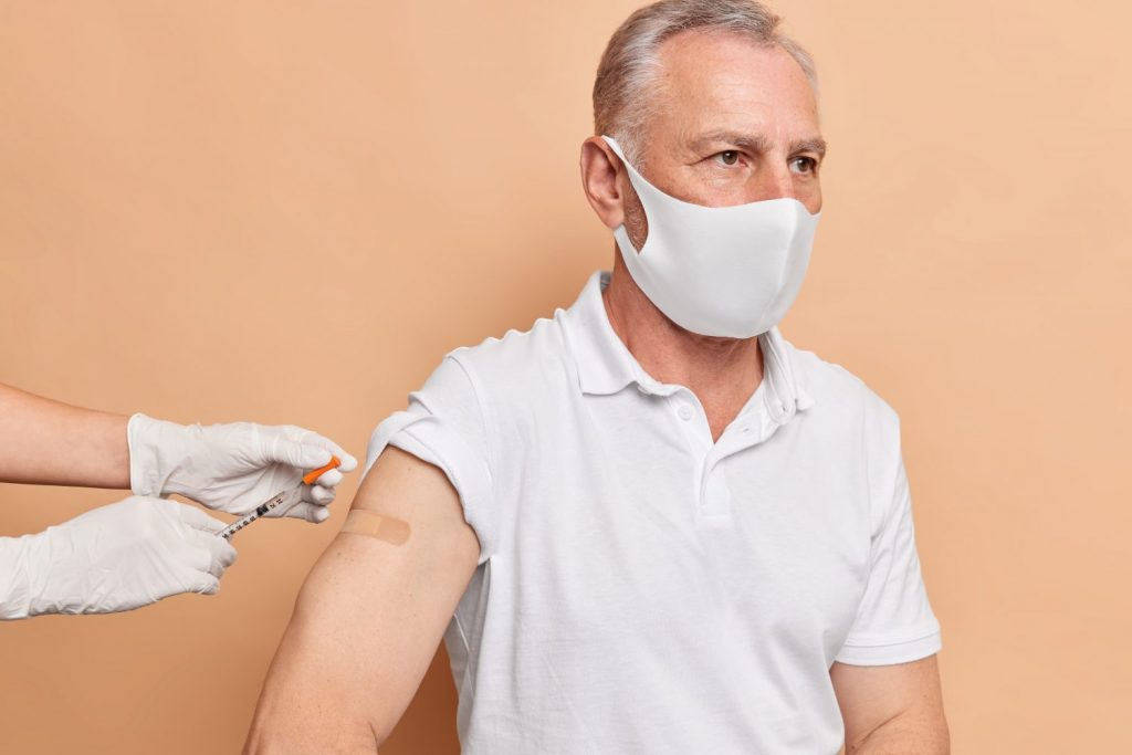 Unrecognizable doctor does vaccination to elderly man. Grey haired male model wears protective face mask fights against coronavirus gets vaccine for disease prevention isolated on beige wall