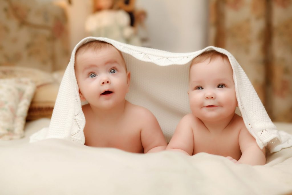 Two twin babies, seven-month smiling girls covered with a white towel in bed on white sheets