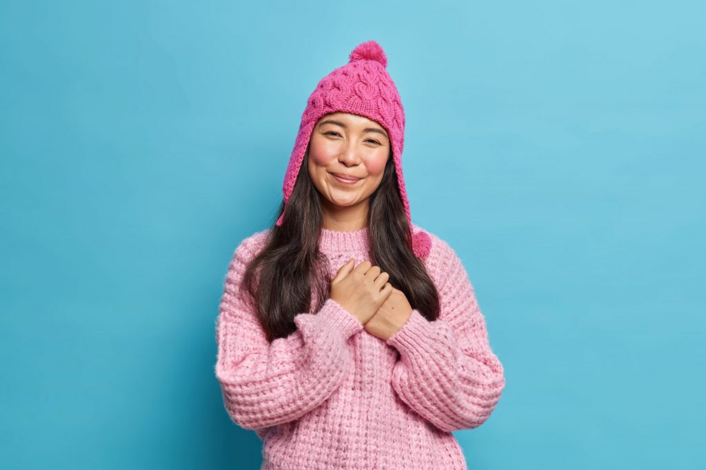 Satisfied good looking Asian woman with dark hair presses hands to heart makes gratitide gesture feels touched wears knitted sweater and hat isolated over blue background. Thank you very much