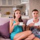 Young Couple with Mugs of Coffee or Tea Sitting on Sofa Together and Watching Television with Blank Expressions