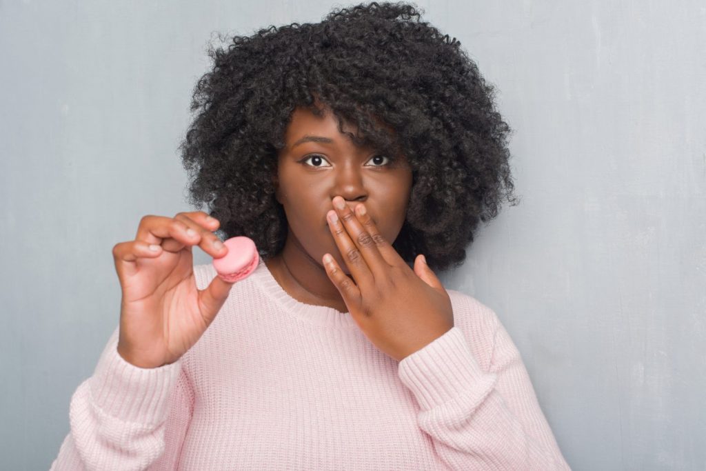 Young african american woman over grey grunge wall eating pink macaron cover mouth with hand shocked with shame for mistake, expression of fear, scared in silence, secret concept