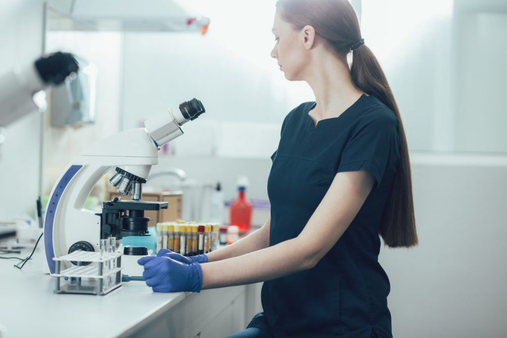 Calm female lab assistant sitting at the table with test tubes and a modern microscope in front of her