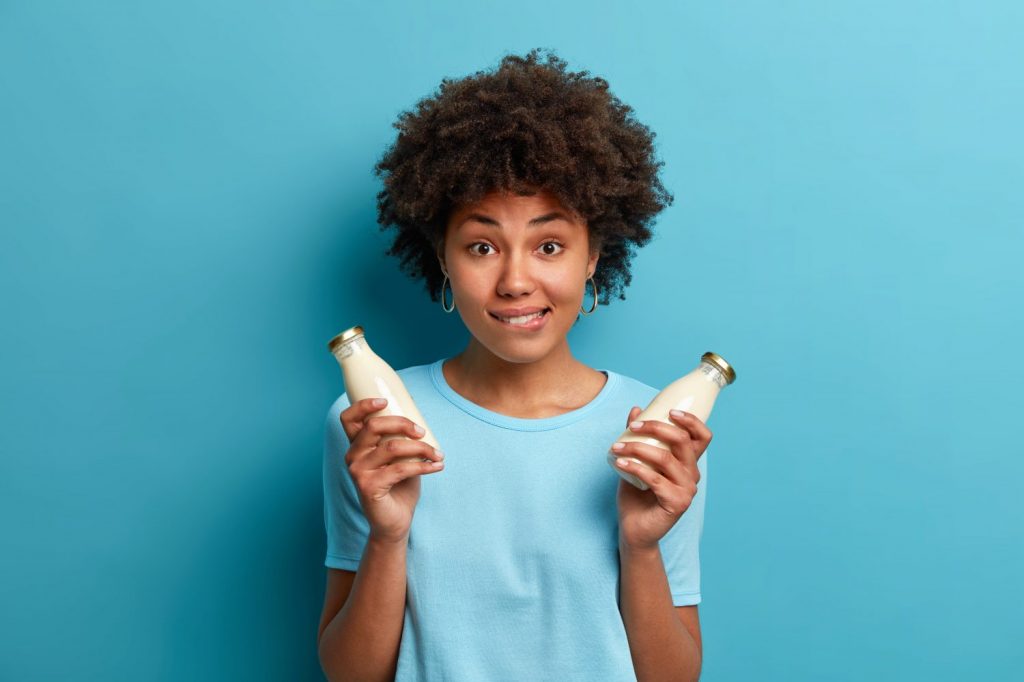 Horizontal shot of woman vegetarian holds two glass bottles of fresh lactose free milk dressed in casual t shirt isolated on blue background. Female model has healthy nutrition poses indoor.