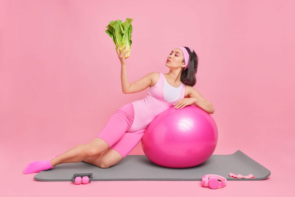 Fitness woman leans on swiss ball holds fresh green vegetable keeps to heathy diet has regular training at home to keep fit isolated over pink background. People proper nutrition and sport concept