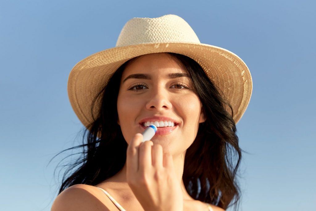 people, summer and swimwear concept - portrait of happy smiling young woman in bikini swimsuit and straw hat applying lip balm on beach