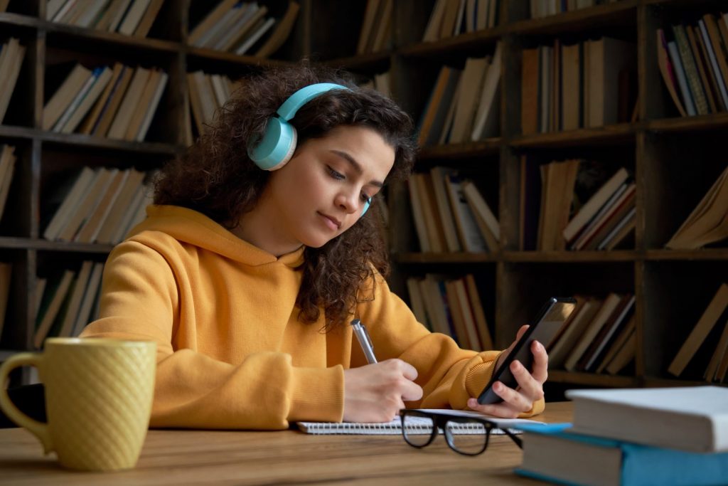 Latin teen girl wear headphones hold phone online learning in mobile app. Hispanic college student using smartphone watching video course, zoom calling making notes in workbook sit in library campus.