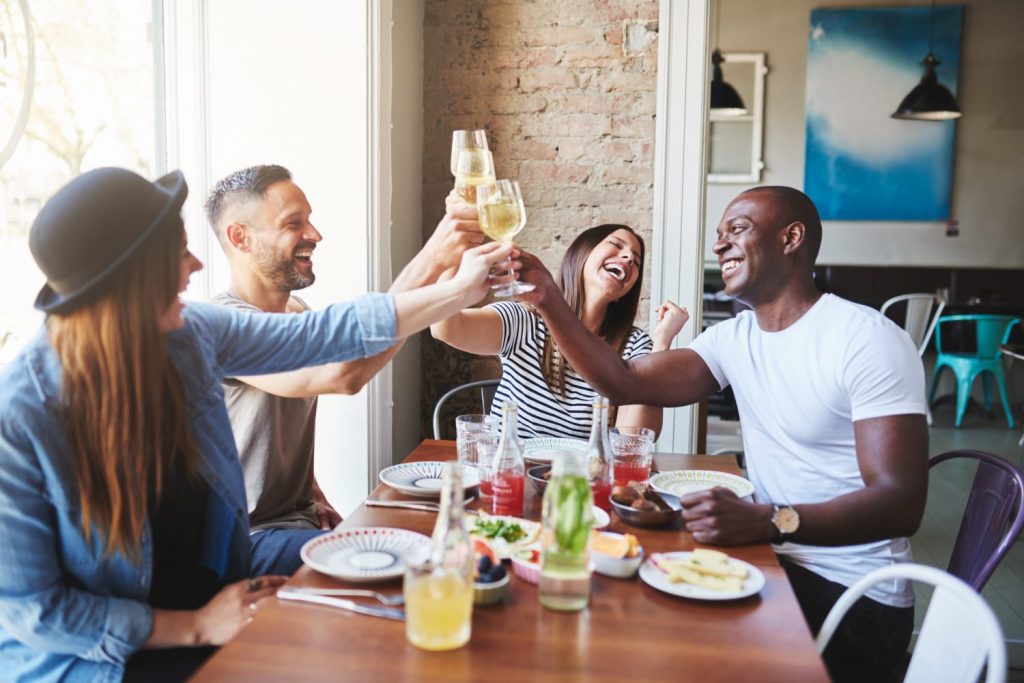 Happy group of diverse male and female young adults in casual clothing toasting drinks together at table in restaurant