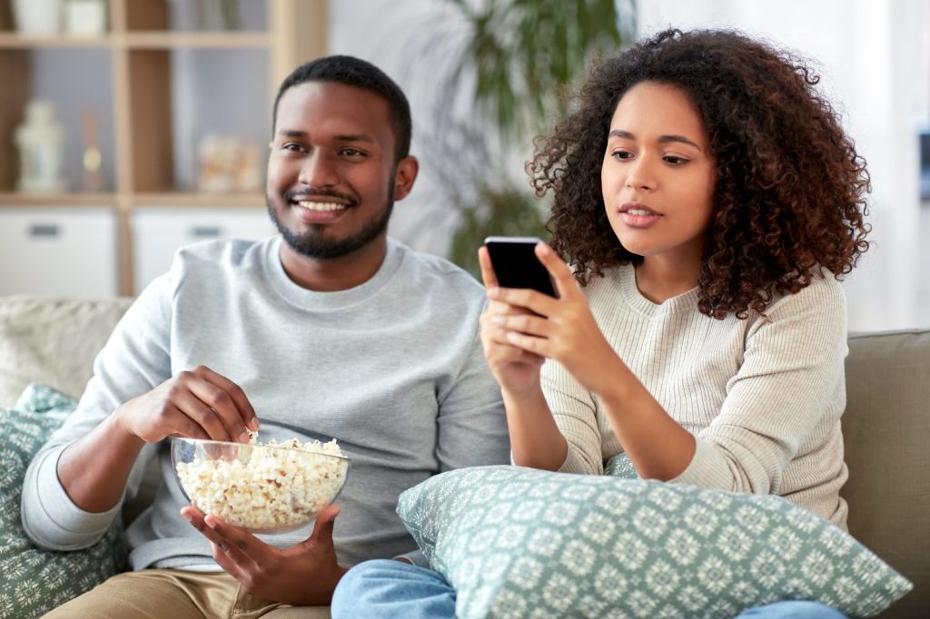 people and leisure concept - african american couple with popcorn and smartphone watching tv at home