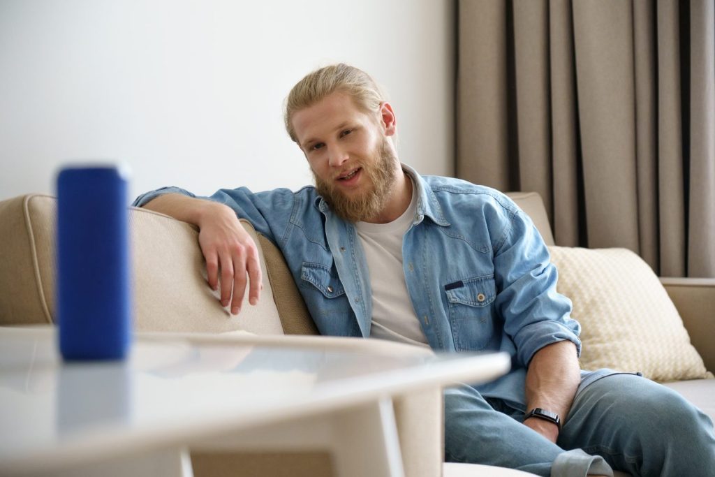 Young man sitting on sofa speaking activating small portable wireless speaker asking virtual voice assistant on table at smart home. Mini bluetooth stereo gadget for sound digital assistance concept.