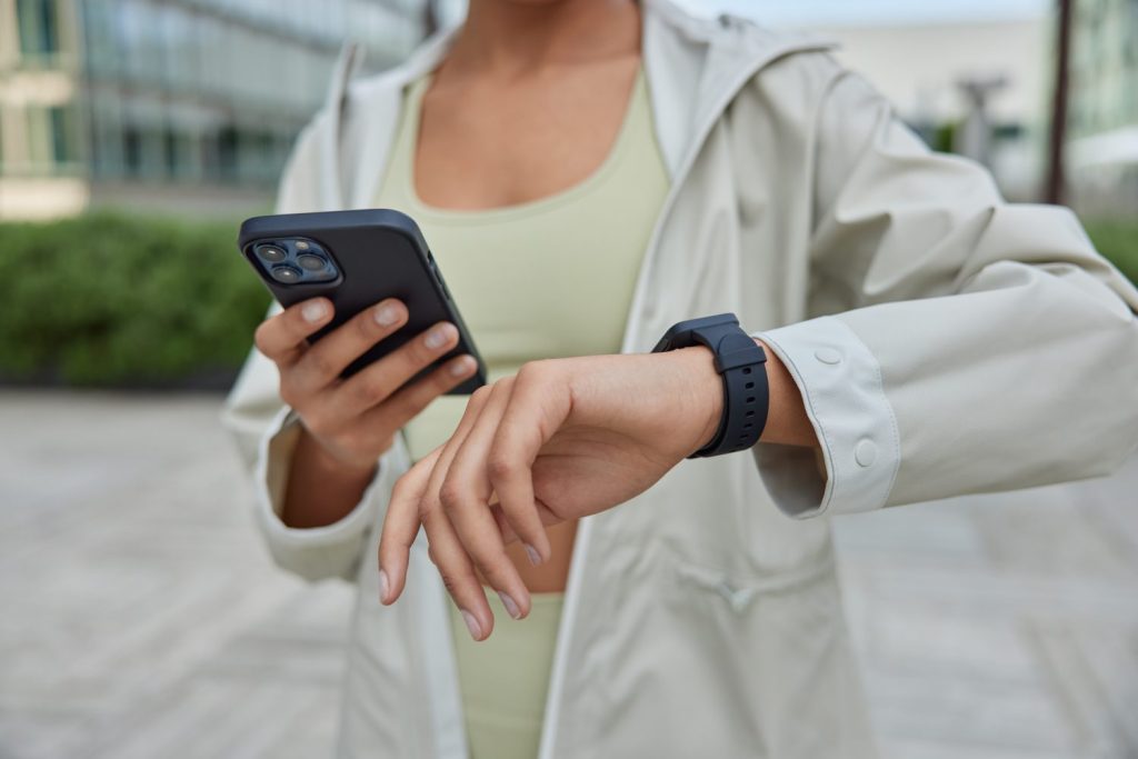 Unknow sportswoman holds digital mobile phone checks fitness tracker results of workout or body vitals on smartwatch dressed in anorak poses outdoors uses modern devices for sport. Cropped shot