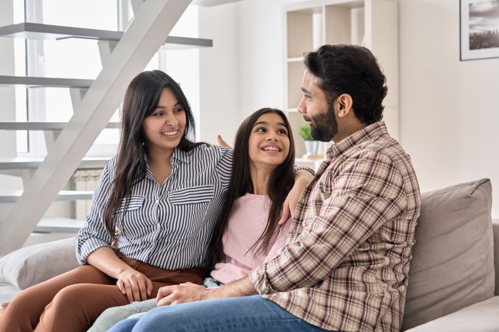 Happy indian family couple with teen child daughter bonding, hugging, talking sitting on couch at home. Smiling husband and wife embracing child spending time together relaxing on sofa in living room