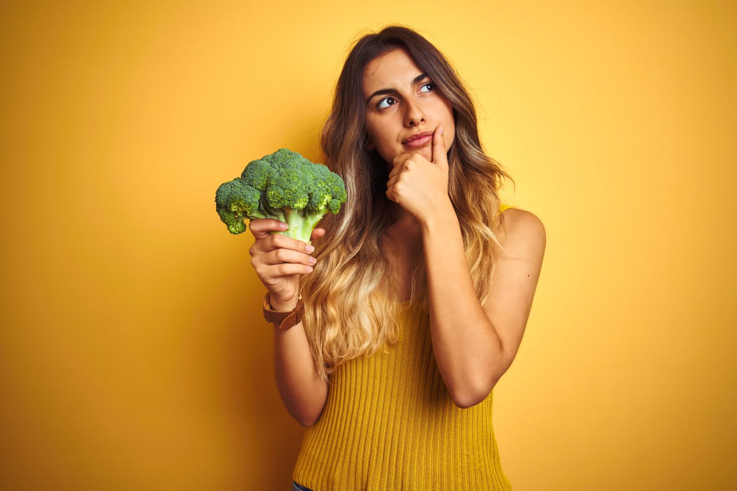 Young beautiful woman eating broccoli over yellow isolated background serious face thinking about question, very confused idea